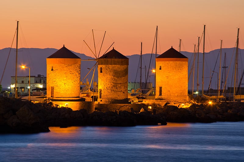 Three stone cylinder buildings near water at sunset with mountain range in background. Fenway Partners Investments image.